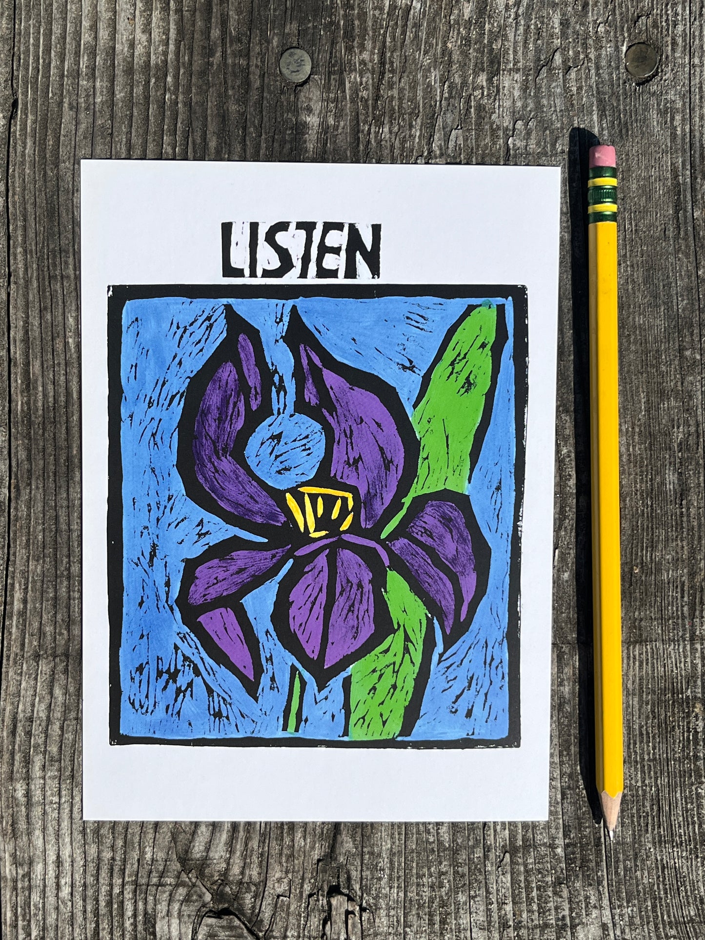 Colorful drawing of a flower with 'LISTEN' on a postcard placed on a wooden surface with a pencil.