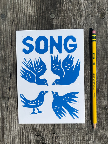 Blue bird illustrations on a white card with 'SONG' text, placed on a wooden surface with a pencil.